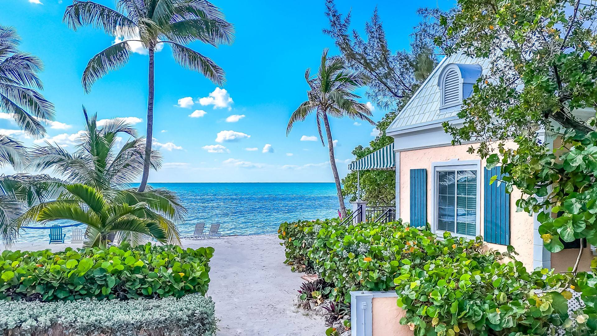 A cottage on a beach with palm trees and two chairs near the ocean.