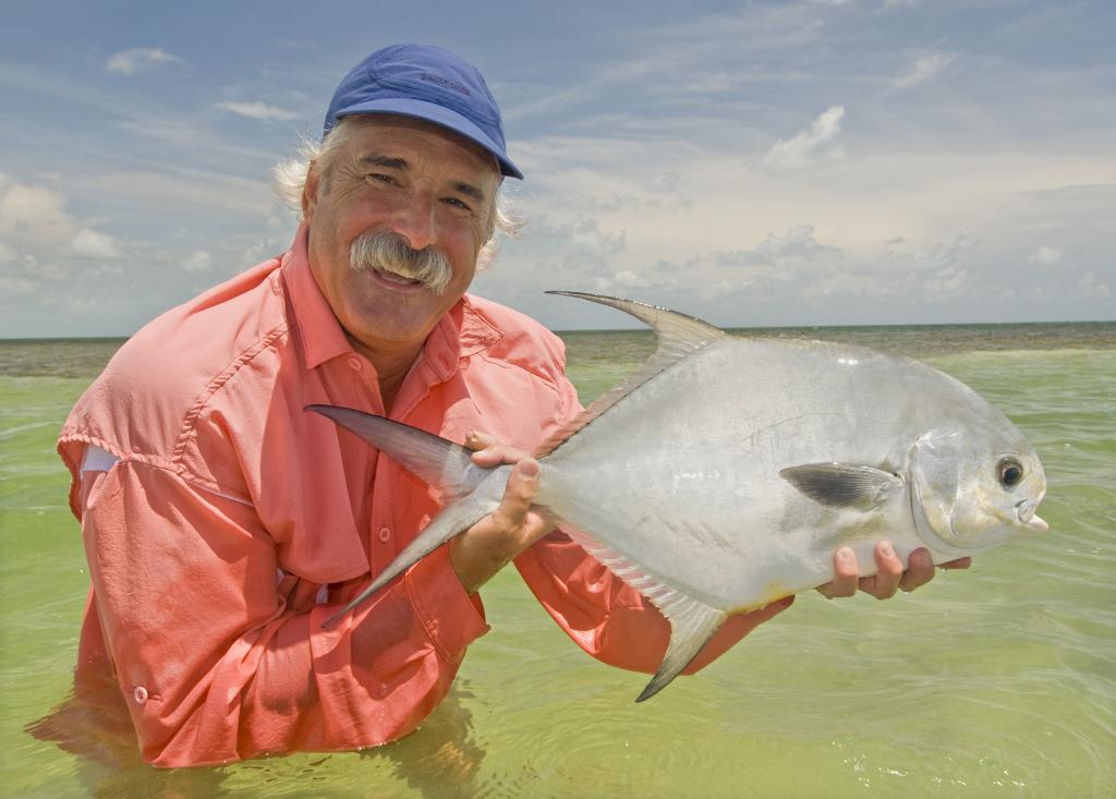 Man in a pink shirt holds a fish in shallow water, smiling under a blue sky.