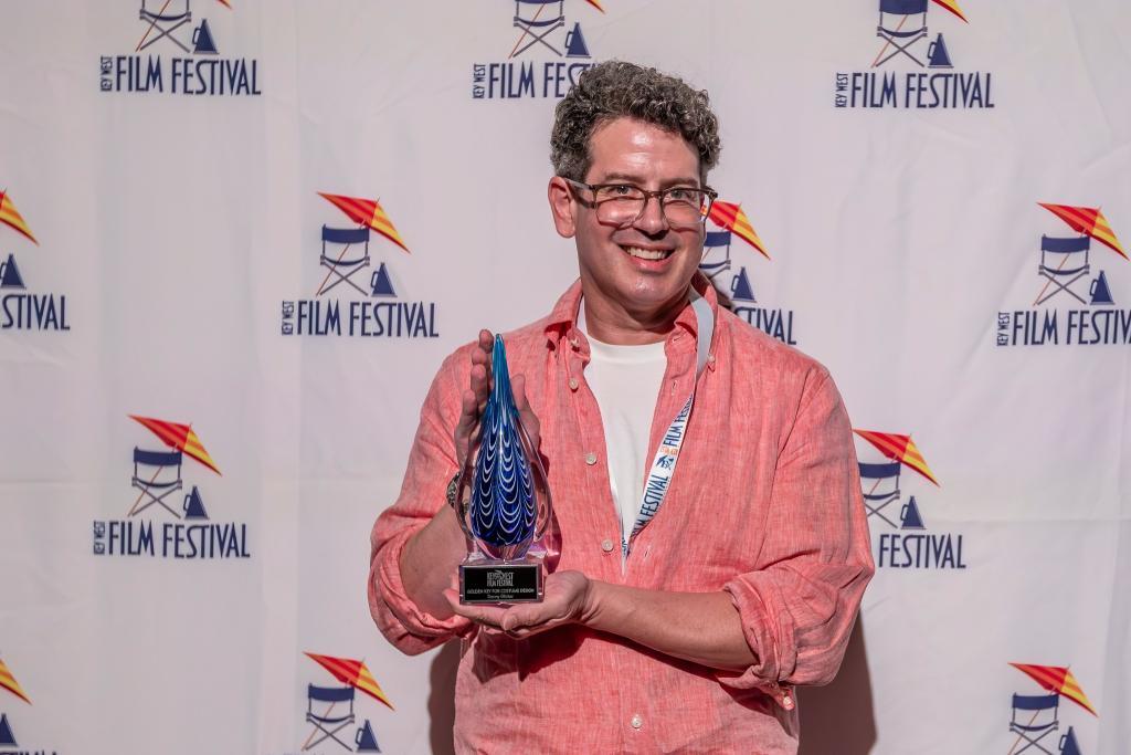 Man in pink shirt holding an award, standing against a festival backdrop.