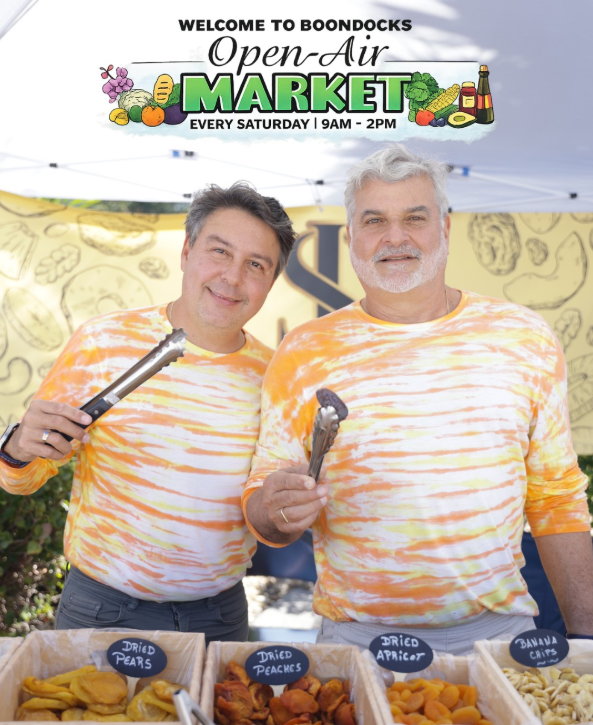 Two men in tie-dye shirts smiling at a market stall with dried fruit.