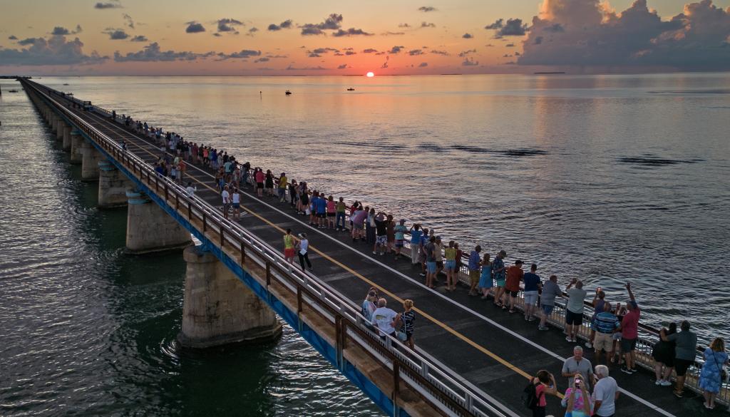 People walking on a bridge over the ocean at sunset.