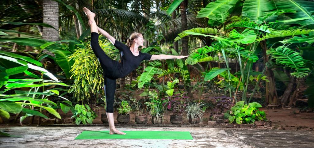 Woman practicing yoga outdoors on a green mat, surrounded by lush tropical plants.