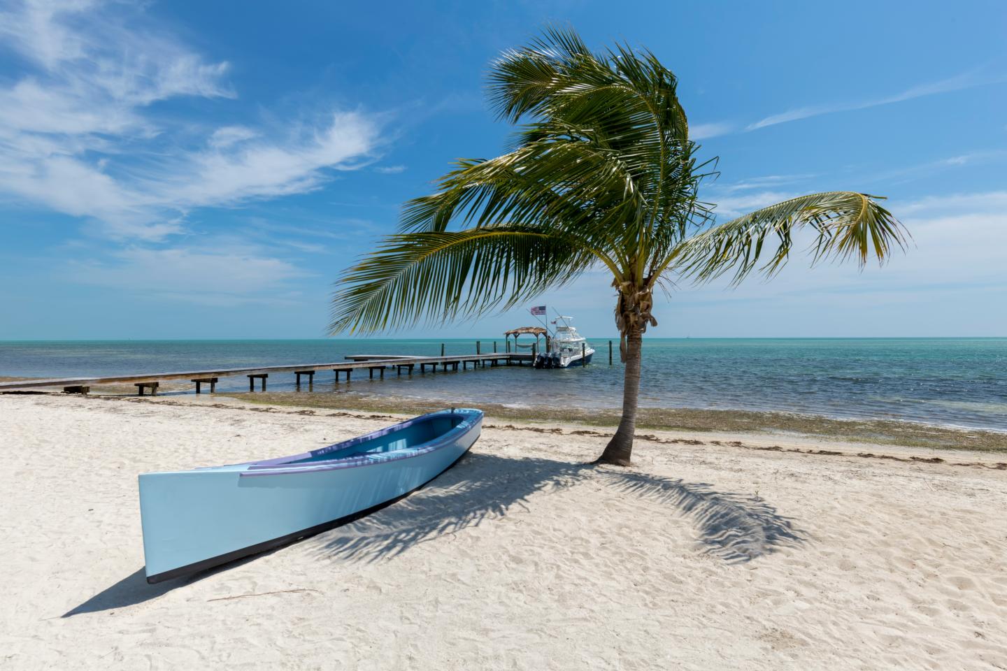 Sandy beach with boat, palm tree, and wooden pier extending into turquoise sea under blue sky.