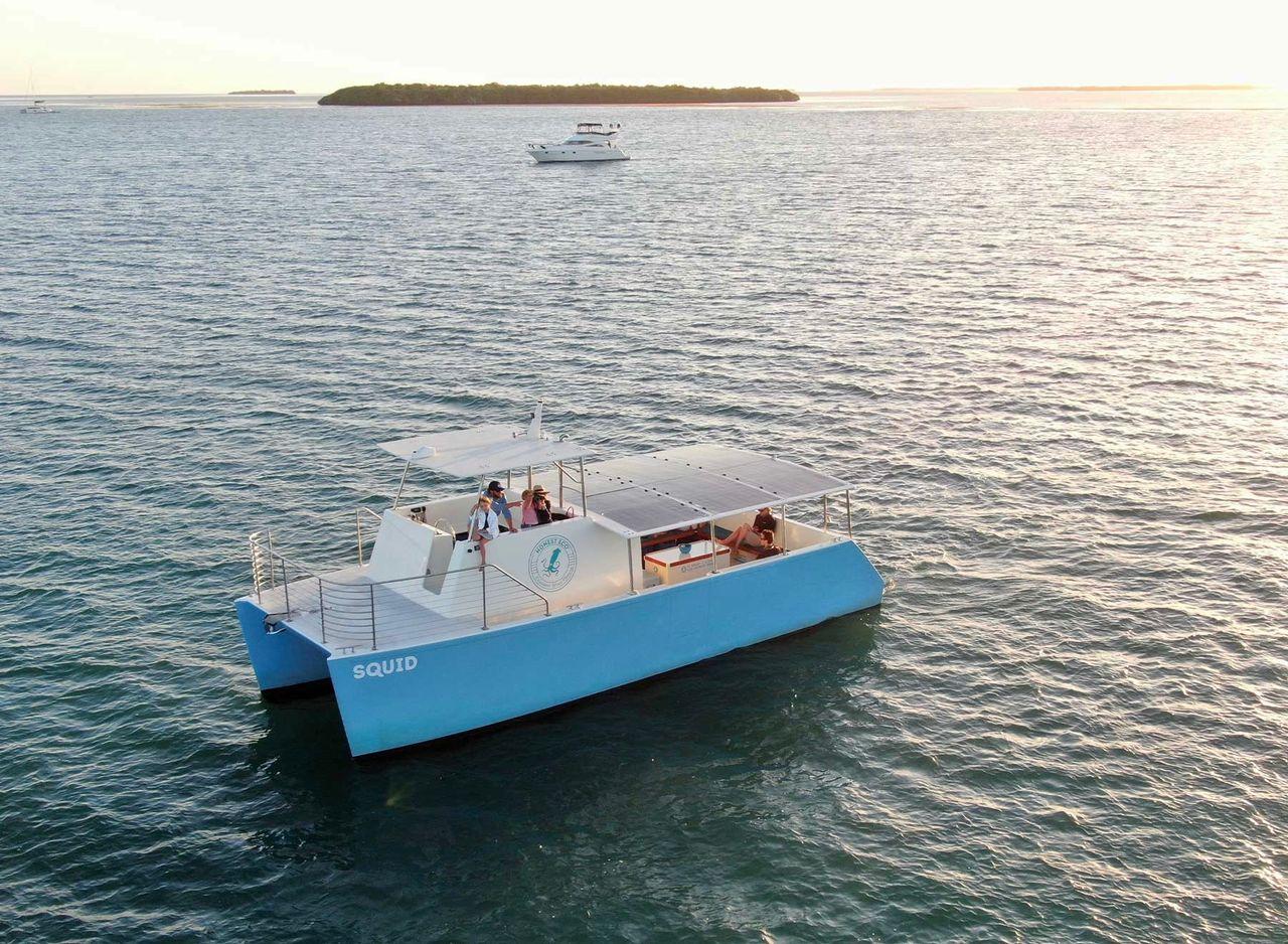 Blue catamaran on calm water during sunset, distant island and another boat in background.