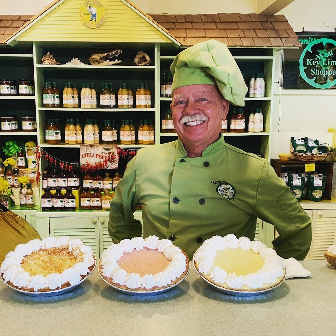 Chef in green uniform smiling behind a counter with three pies.