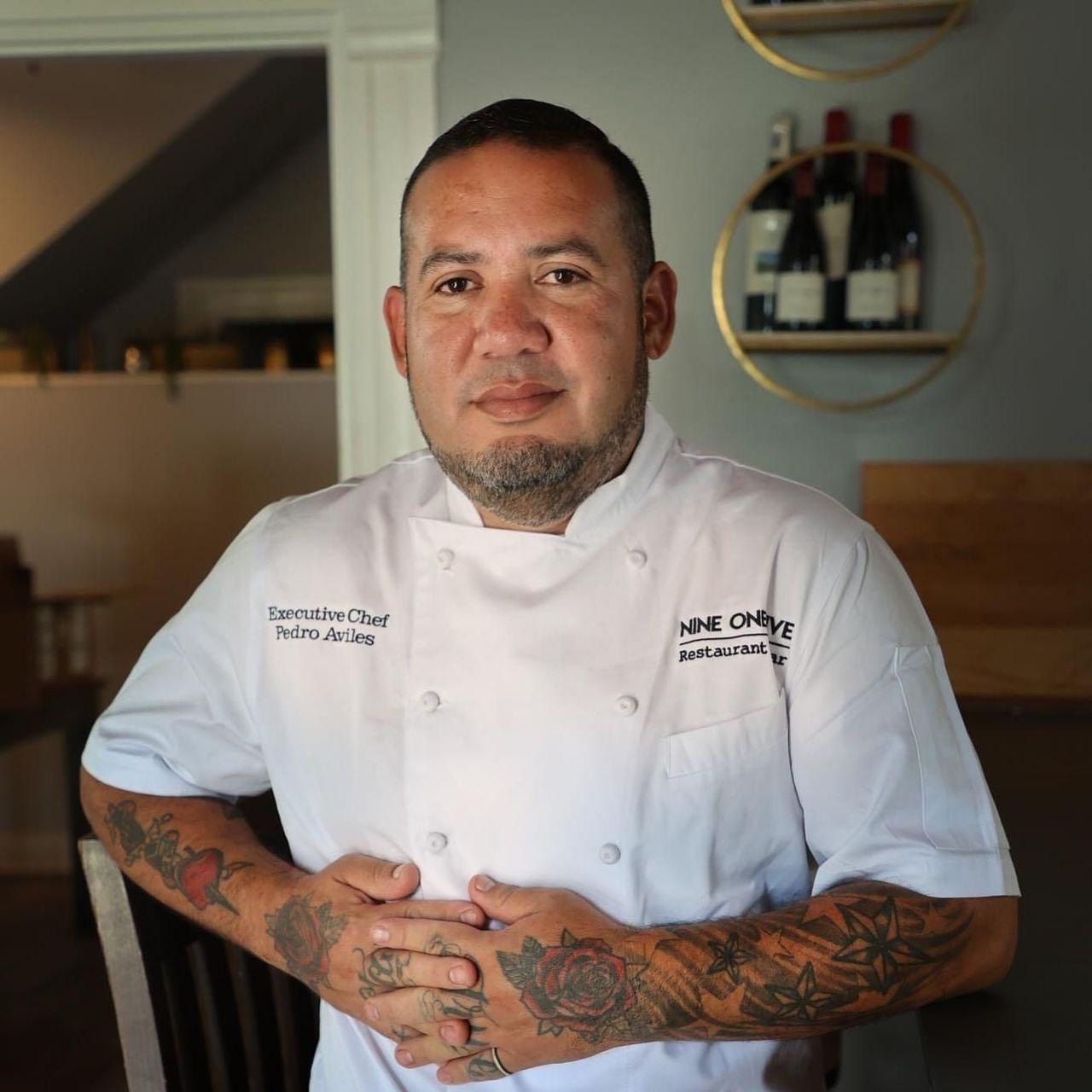 Chef in a uniform with tattoos, standing indoors, hands clasped.