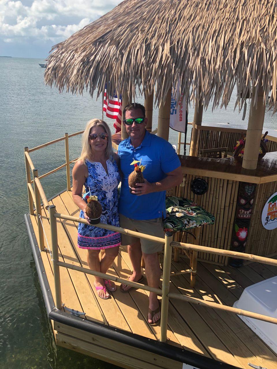 Couple on a tiki bar boat holding drinks, sunny weather.