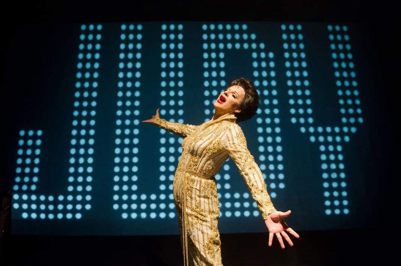 Performer in a gold outfit poses exuberantly on stage, "JURY" lit in background.