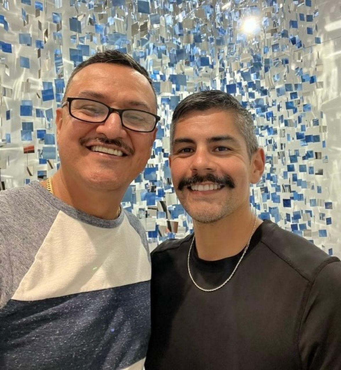 Two smiling men with mustaches in front of a silver and blue decorative backdrop.