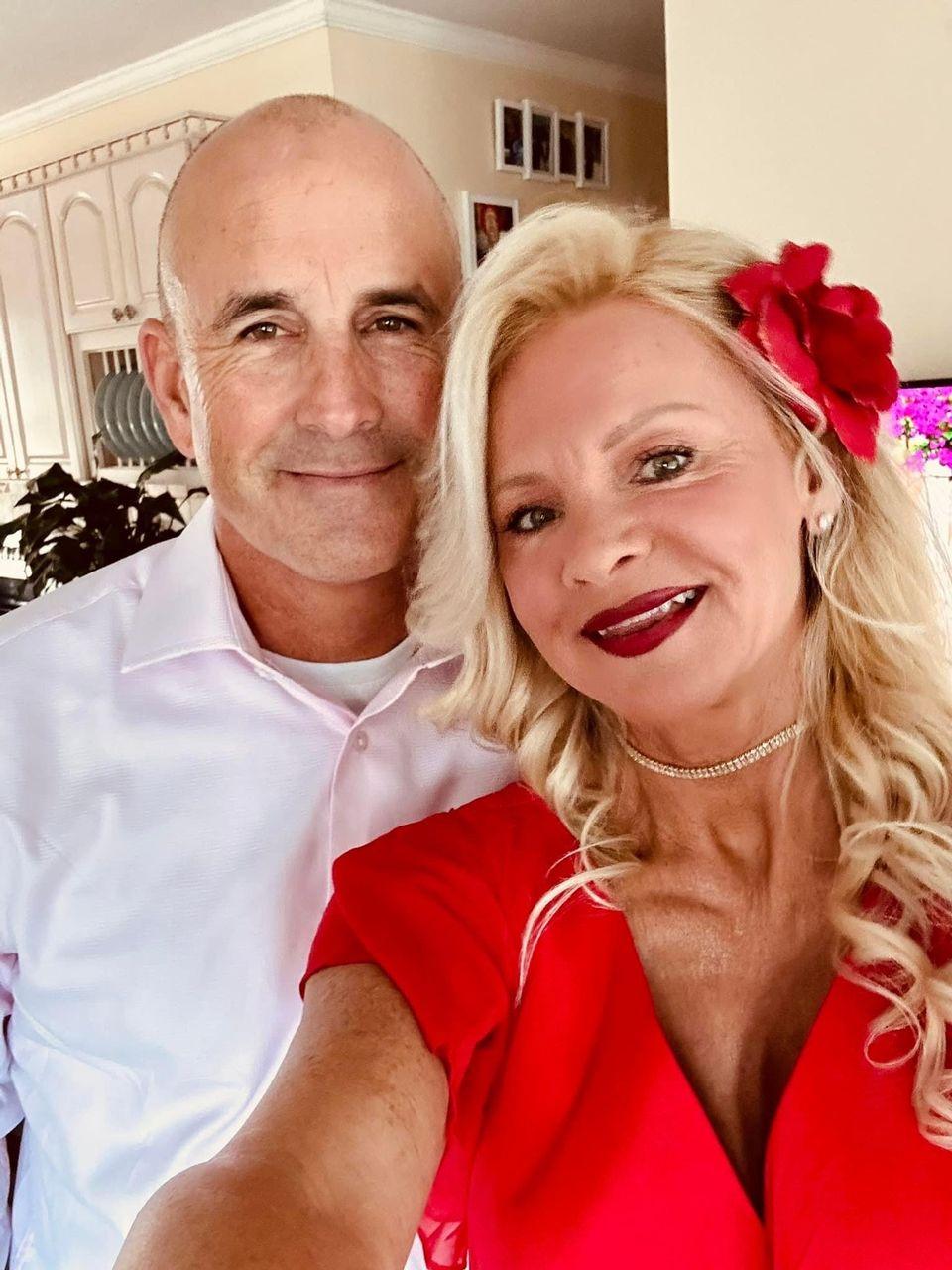 Smiling couple posing indoors, woman in red dress with flower, man in white shirt.