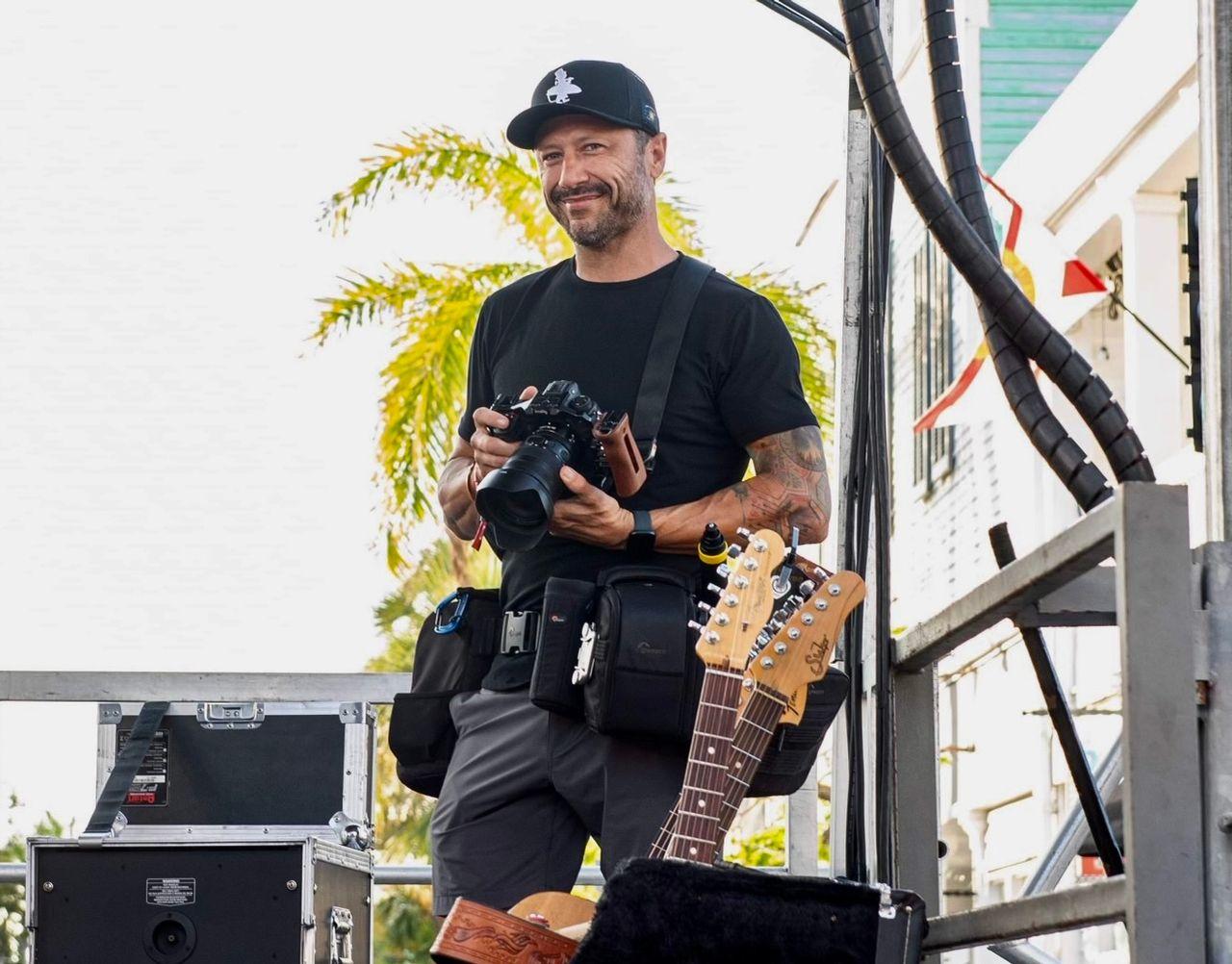Man with a camera on stage, guitars in foreground, palm tree behind.