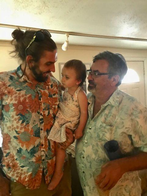 Three people smiling indoors, two men in floral shirts holding a toddler.