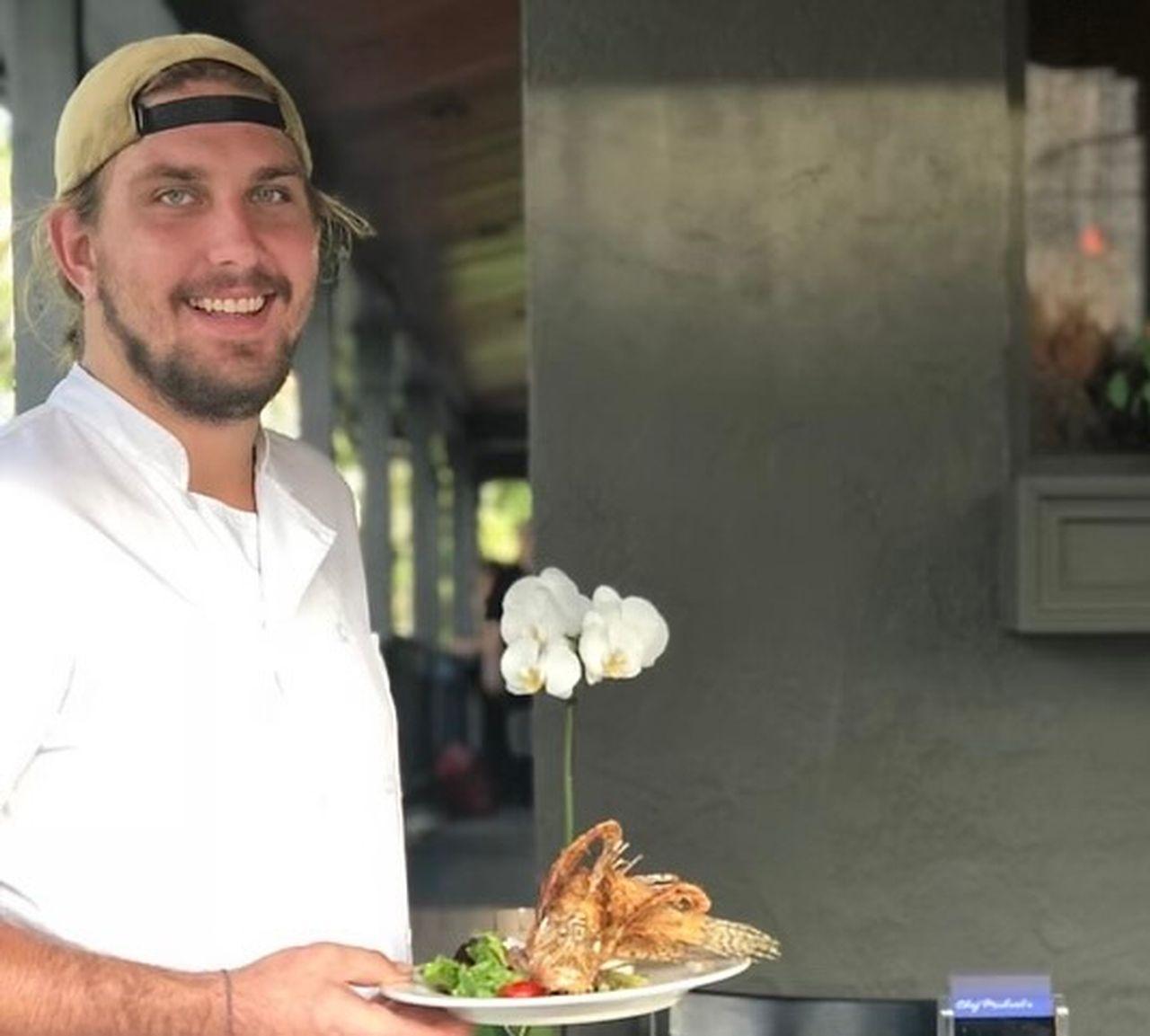 Smiling chef holding a plate of food in an outdoor setting.