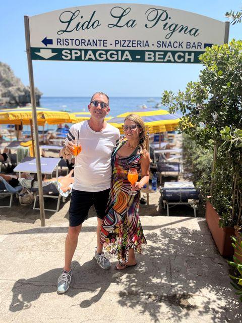 Couple smiling, holding drinks, in front of beachside restaurant sign.