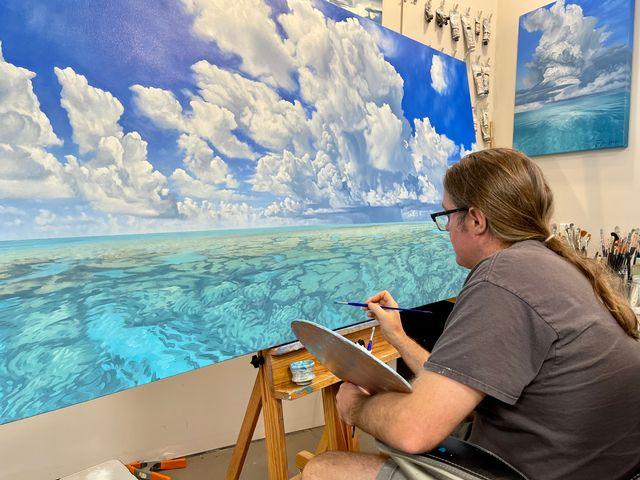 Artist painting a seascape with clouds on a large canvas.