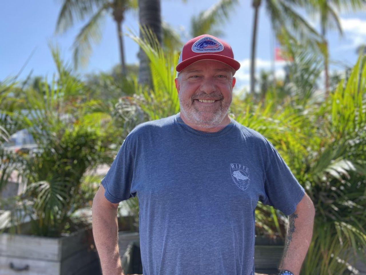 Man in a red cap and blue shirt, smiling outdoors with palm trees in the background.