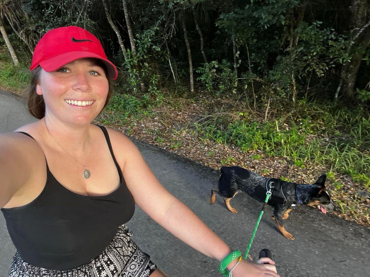 Woman biking with a dog on a wooded path, wearing a red cap and smiling.