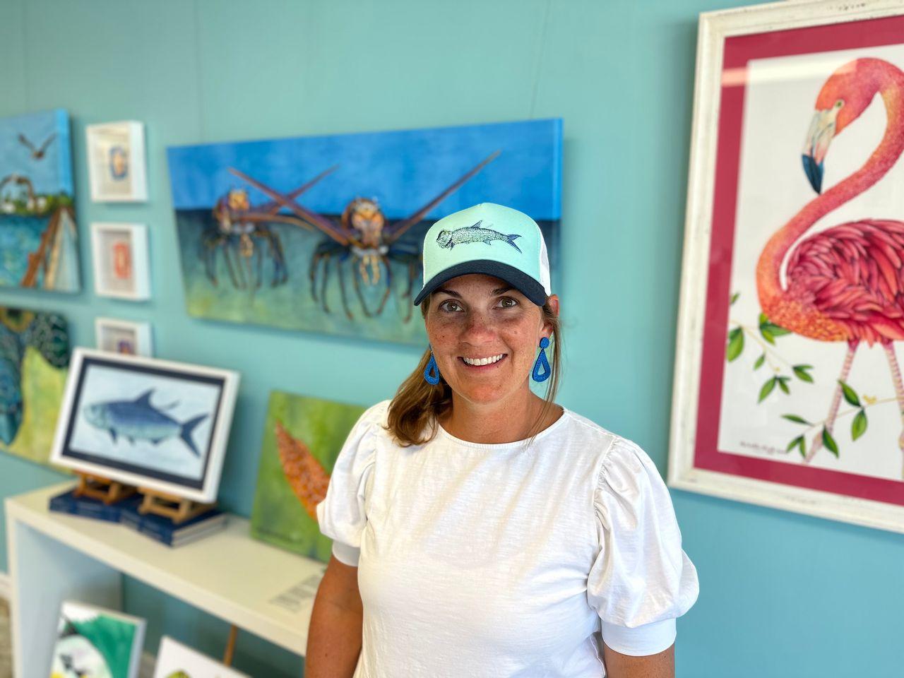 Woman in a gallery, smiling, wearing a turquoise cap and white blouse, colorful art on walls.