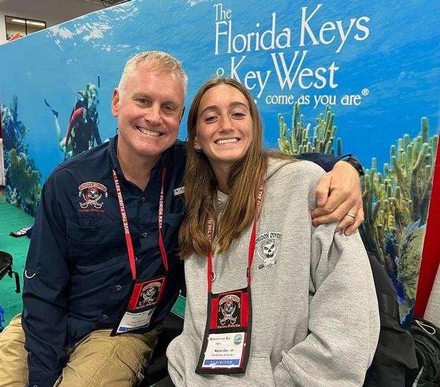 Man and woman smiling, seated in front of a Florida Keys promotional backdrop.