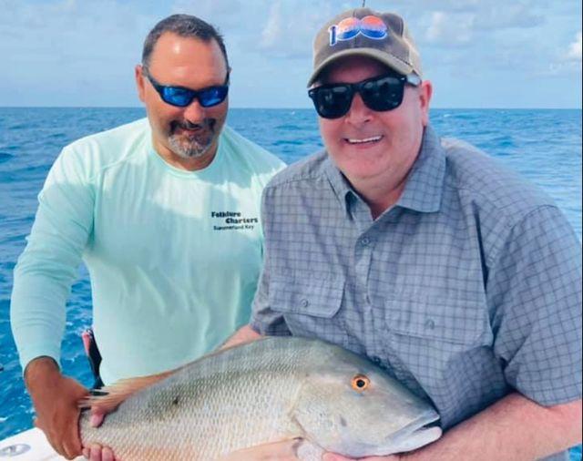 Two men on a boat holding a large fish, smiling under a clear blue sky.