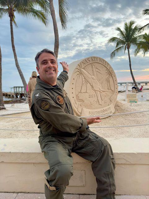 Man in uniform poses with sand sculpture, palm trees in background.