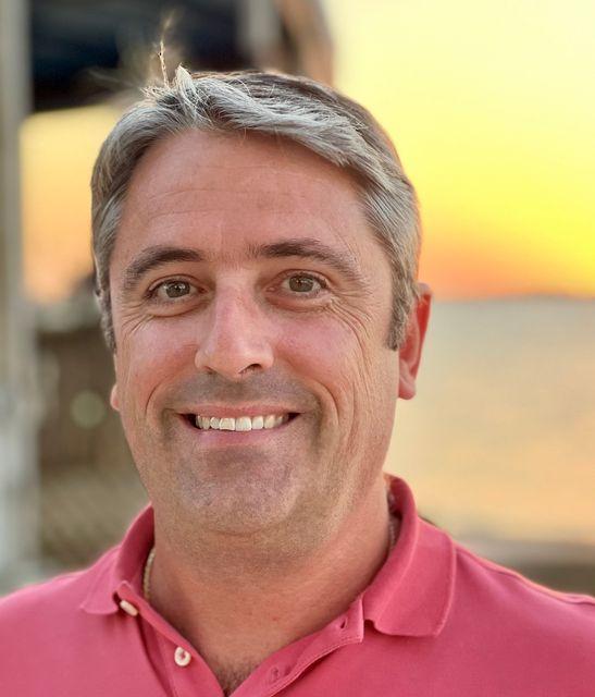 Smiling man in a pink shirt with sunset in the background.