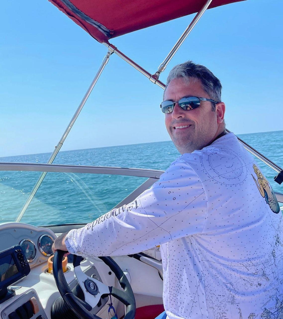 Man steering a boat on a sunny day, wearing sunglasses and a white shirt.