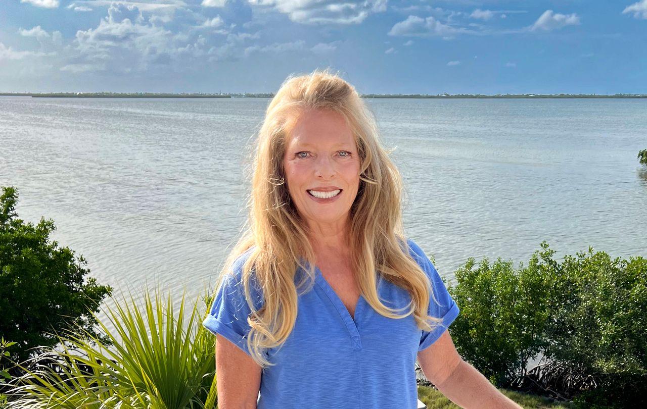 Smiling woman in blue top by calm water and greenery under a blue sky.