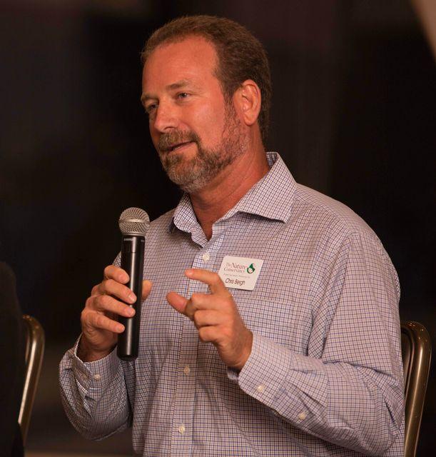 Man speaking into a microphone, wearing a checked shirt.