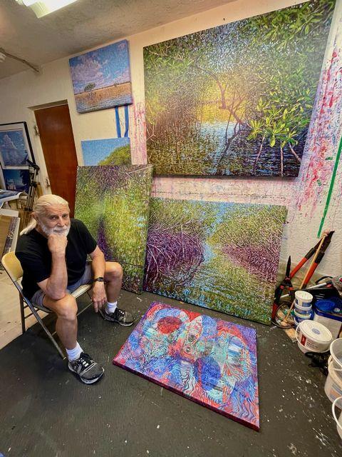 Artist sitting in studio with colorful abstract paintings on walls and floor.