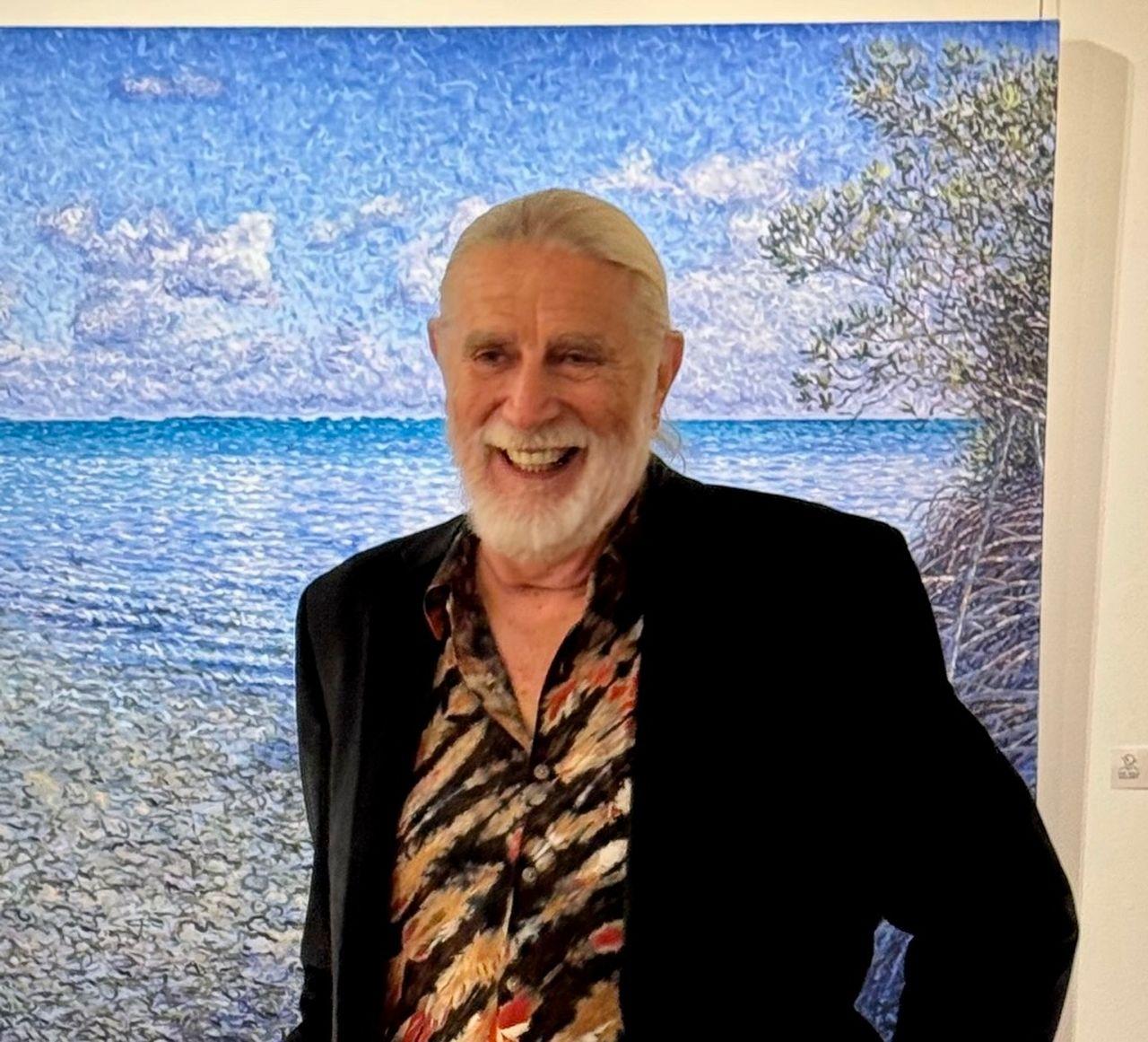 Man smiling in front of a vibrant seaside painting.