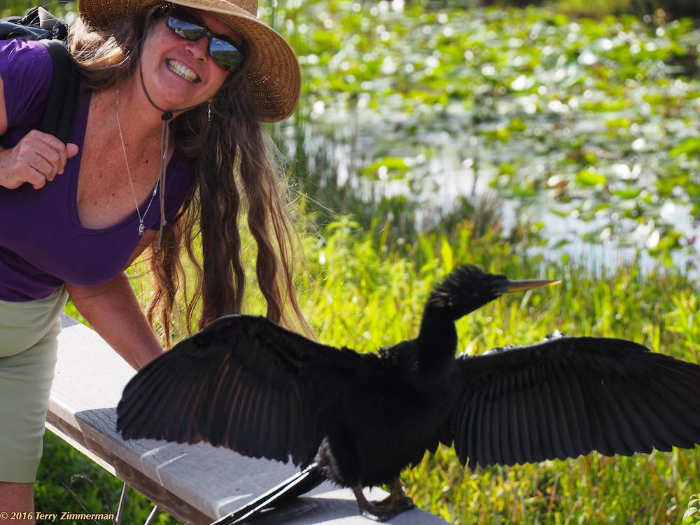 Woman smiling with a hat, next to a bird with open wings by a pond.