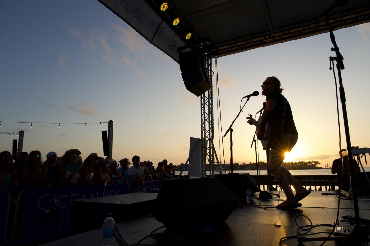 Musician silhouetted against a sunset, playing guitar on stage to a crowd.