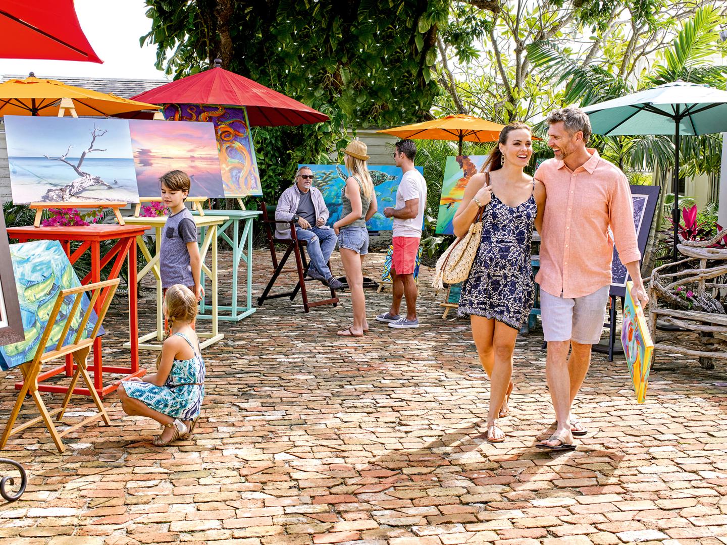 Outdoor art market with people browsing colorful paintings under umbrellas.