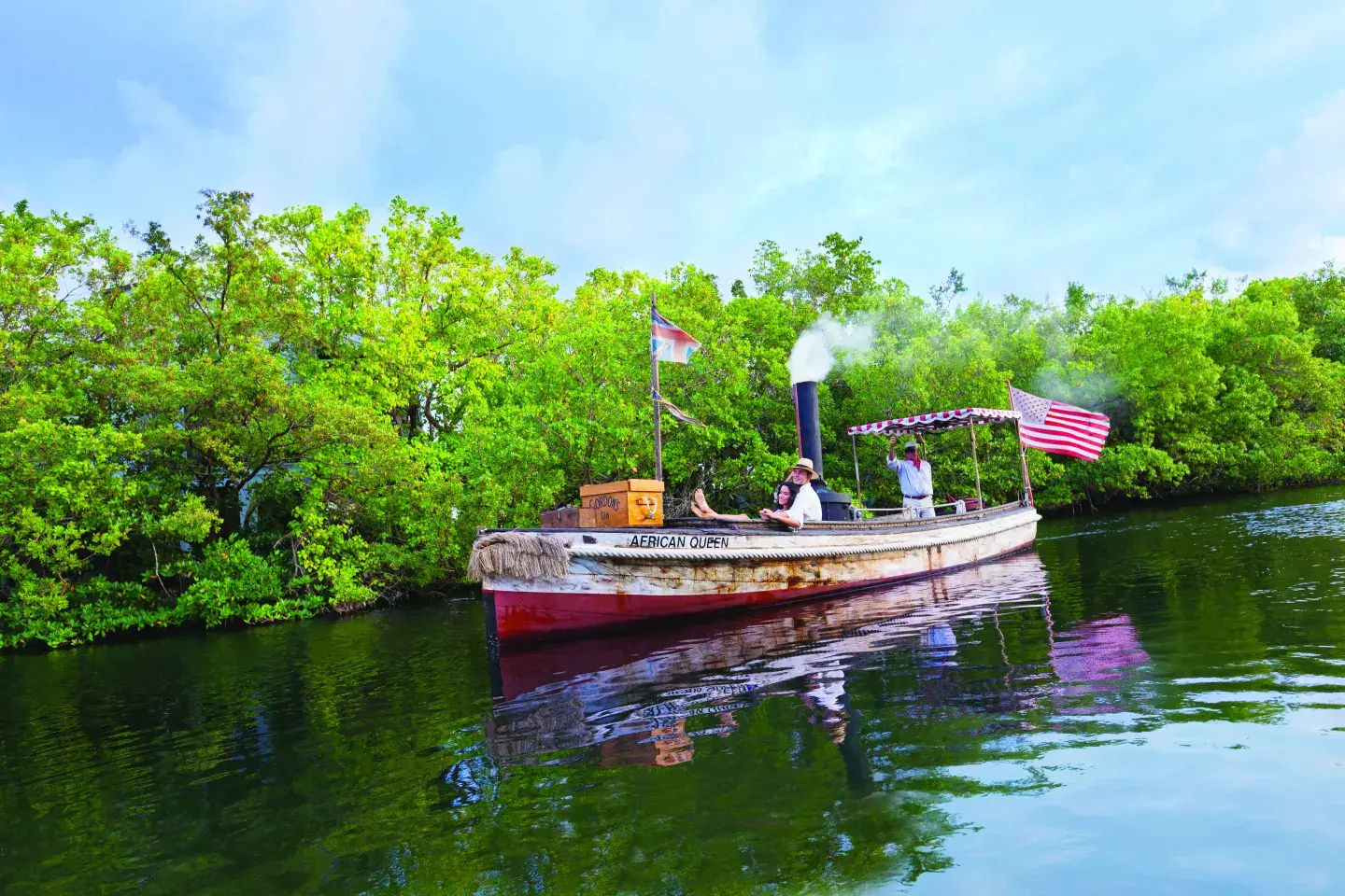 Old steam-powered boat on a lush, green river under a bright, cloudy sky.
