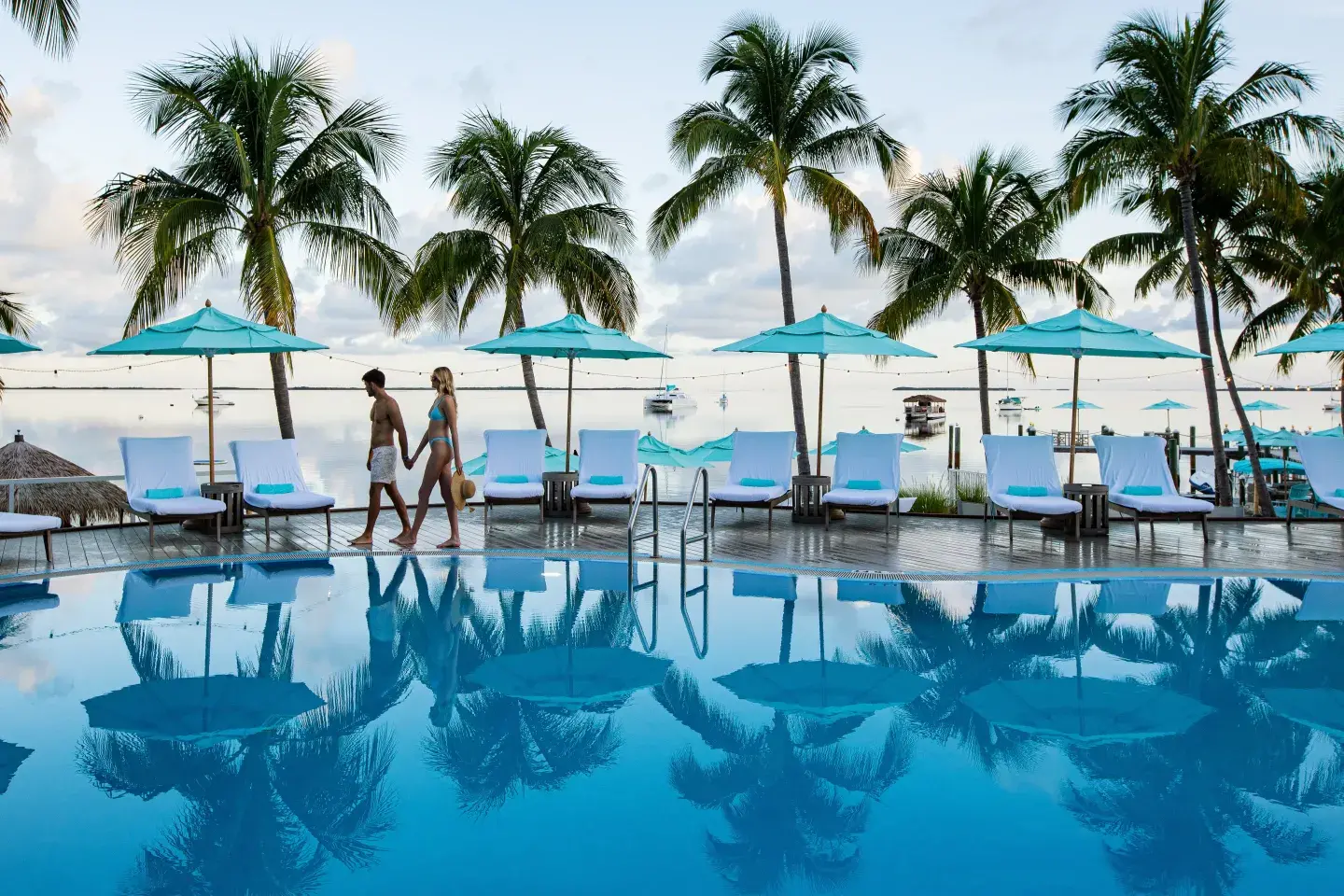 Poolside with palm trees, umbrellas, and two people walking near the water.
