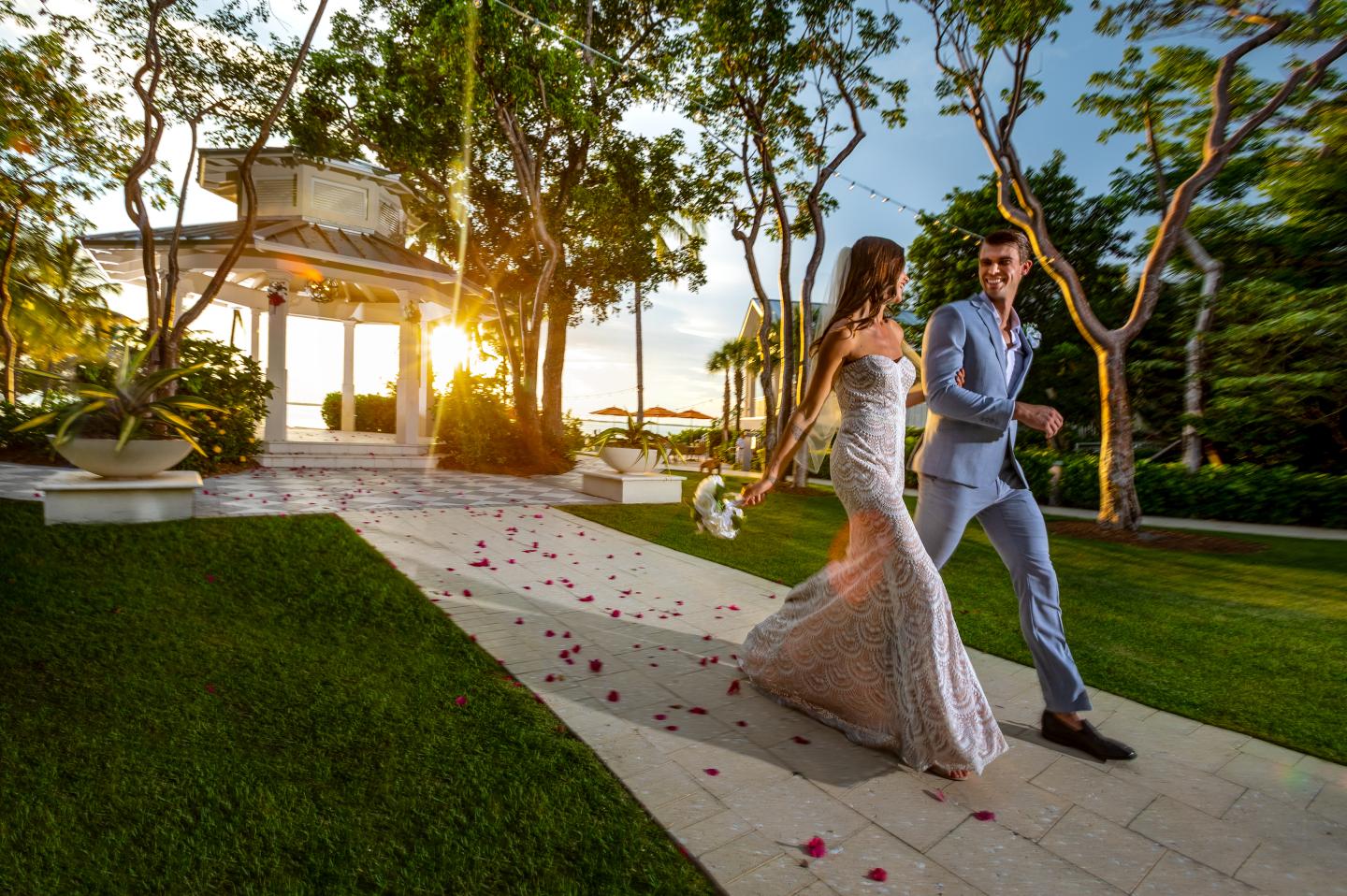 Couple in wedding attire walking on a garden path at sunset.