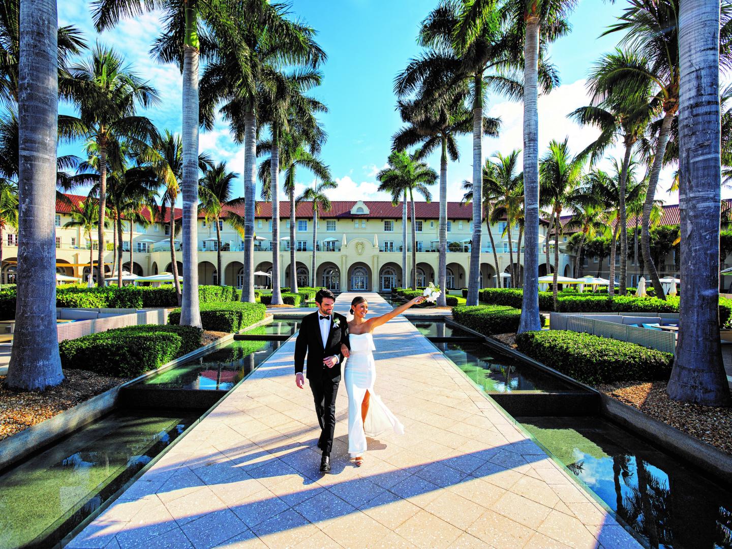 Bride and groom walking on a sunny path lined with palm trees.