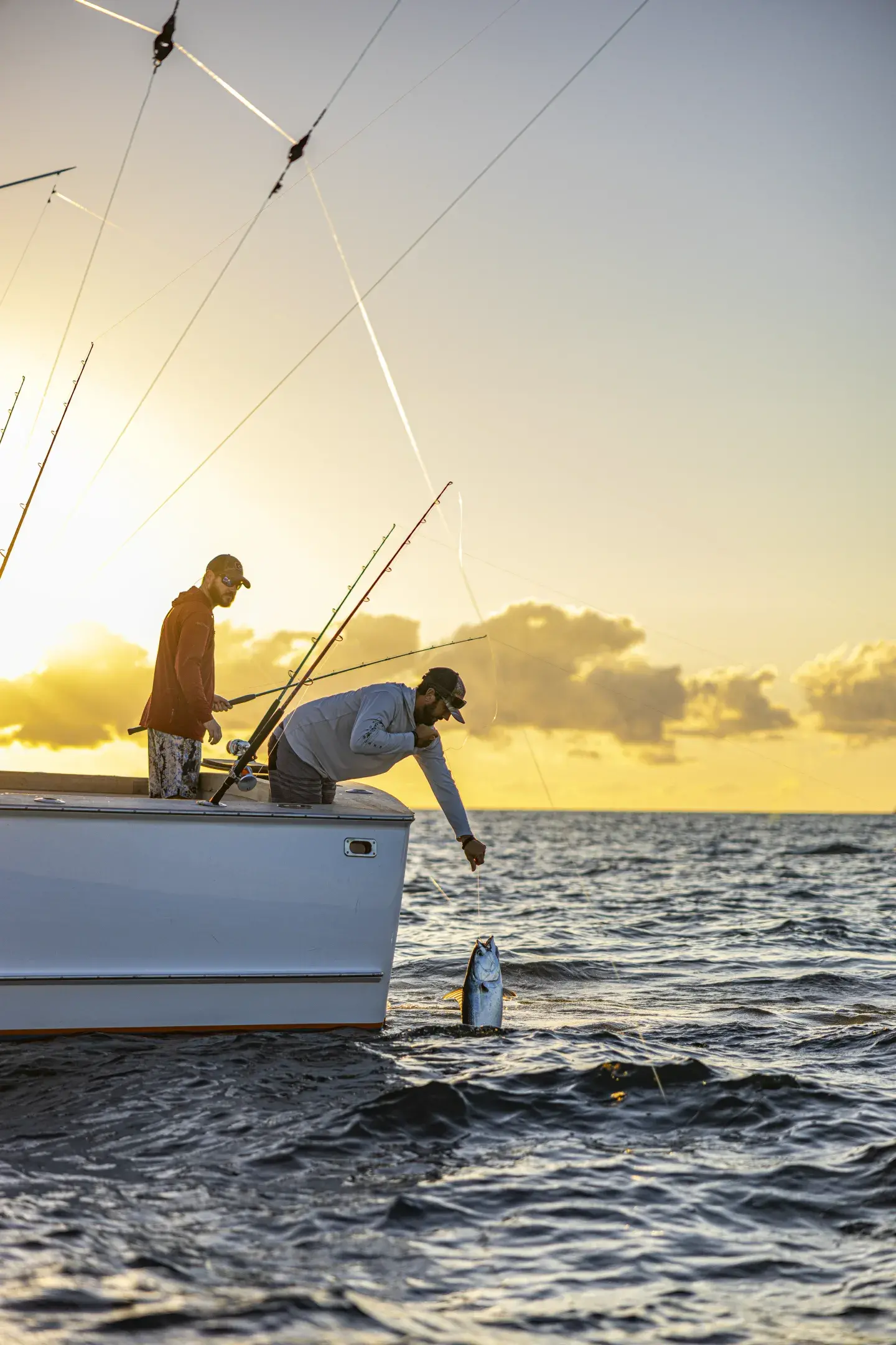 Two people fishing on a boat at sunset, one reaching for a fish.