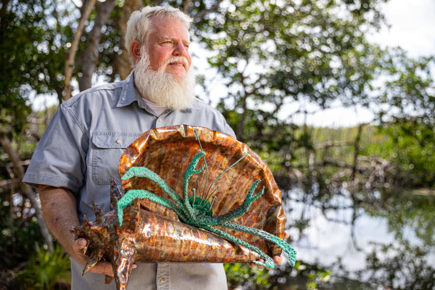 Bearded man holding large shell art outdoors by a swamp.