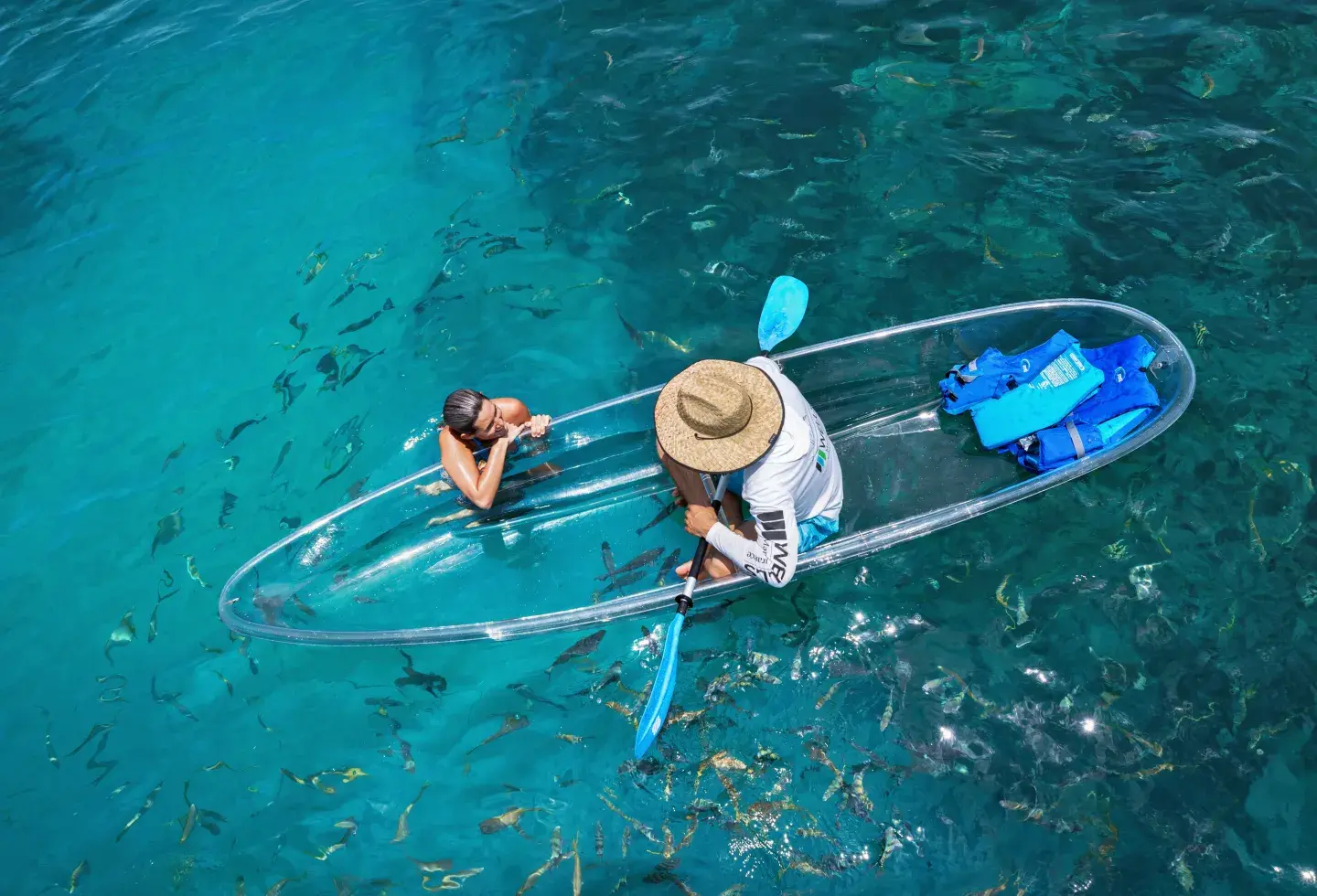 Transparent kayak on clear blue water with fish below; two people inside.