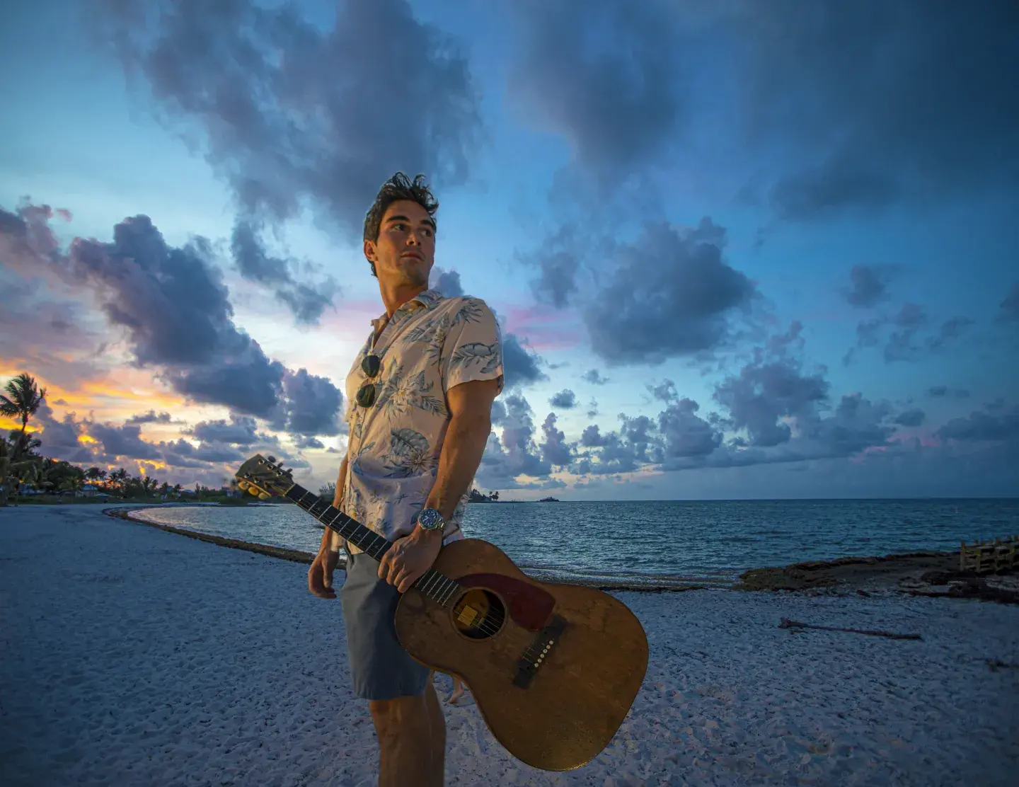 Man holding guitar on a beach at sunset, clouds overhead.