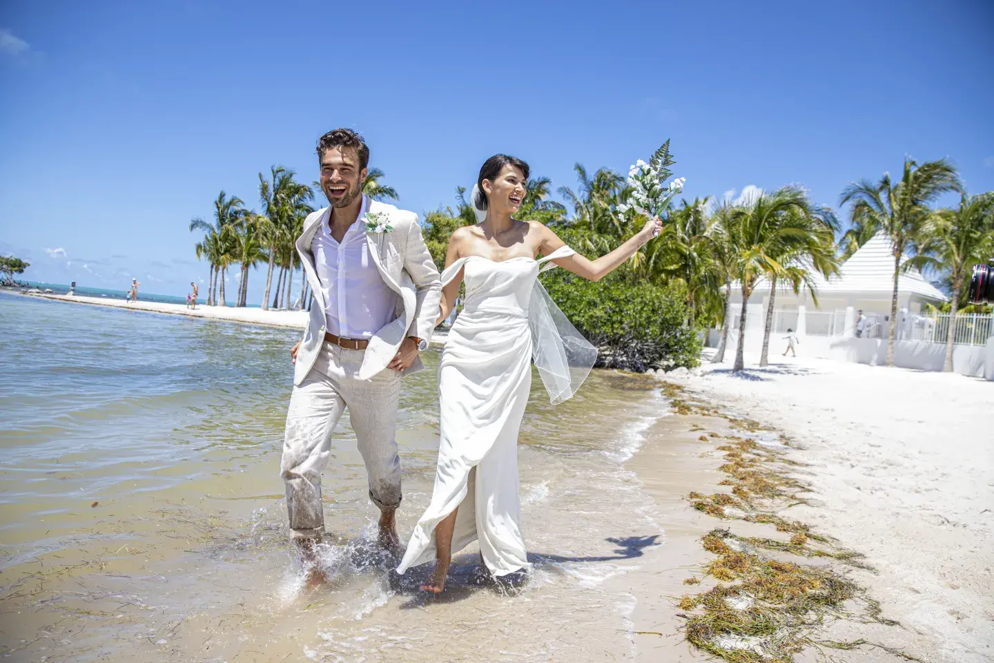 Couple in wedding attire walking on a sunny beach with palm trees.
