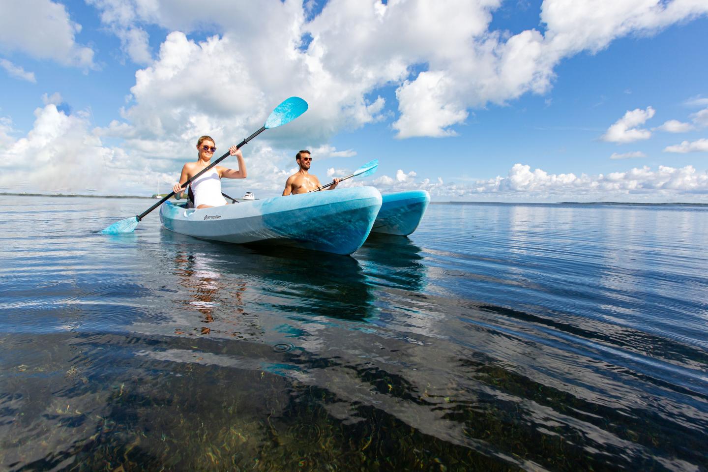 Two people kayaking on a calm lake under a blue sky with clouds.