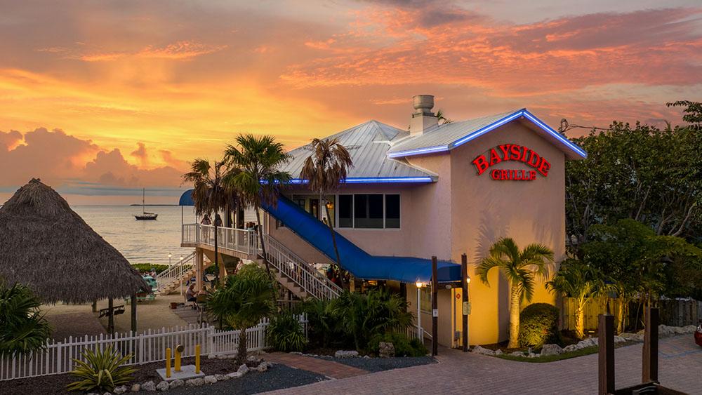 Beachfront restaurant at sunset, palm trees and ocean in the background.
