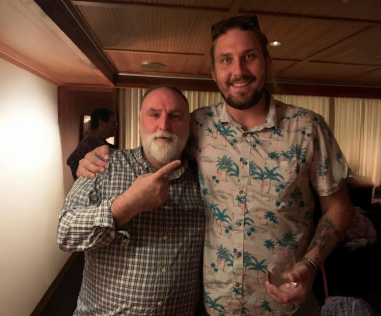 Two men smiling, posing in a warmly lit room; one points, the other holds a drink.
