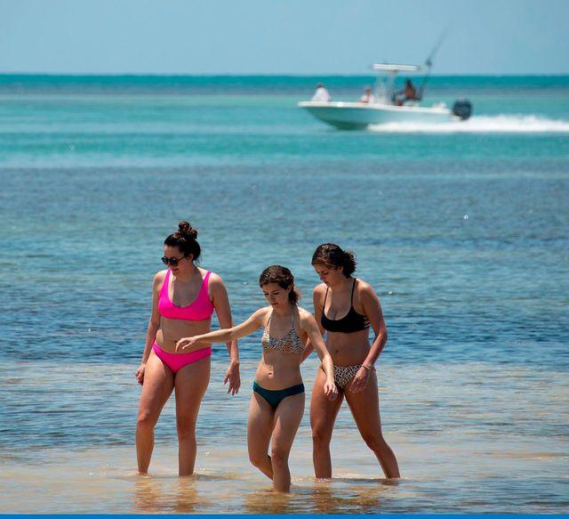Three women walking in shallow ocean water with a boat in the distance.
