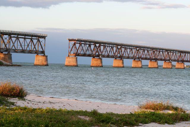 Old railway bridge over calm ocean water with a grassy shore.