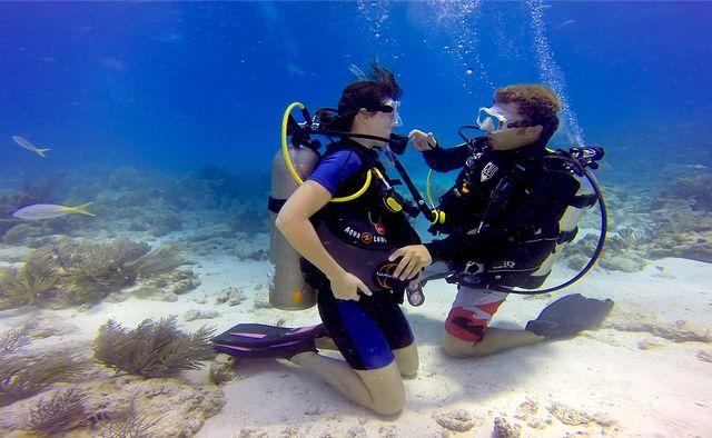Two scuba divers kneeling on the ocean floor, surrounded by coral and clear blue water.