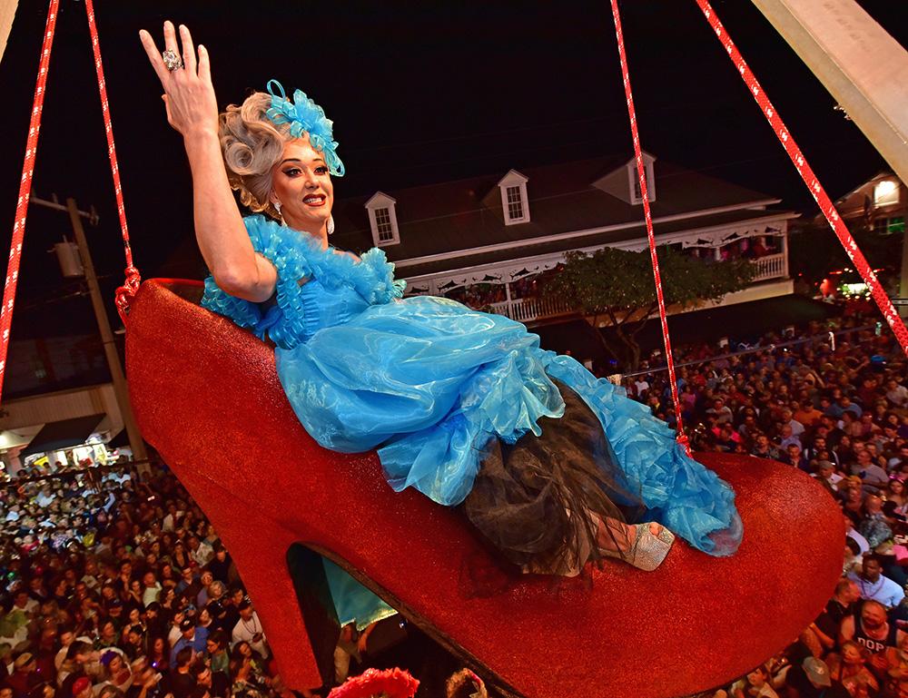 Drag queen in blue dress sits on giant red shoe above a lively crowd at night.